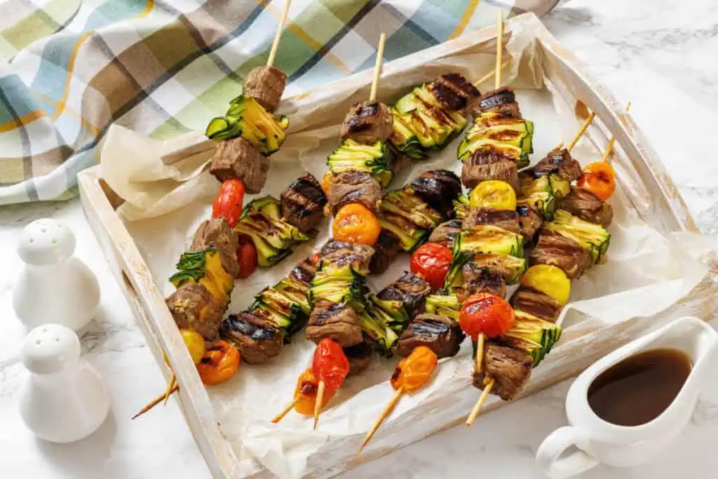 Grilled skewers with beef, zucchini, and cherry tomatoes on a parchment-lined tray. A sauce pitcher and salt and pepper shakers are nearby.