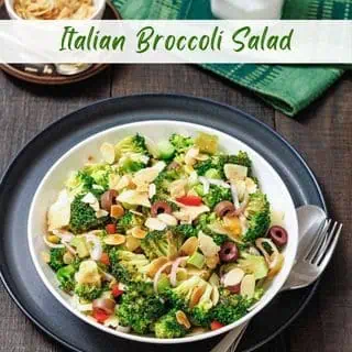 A bowl of broccoli salad with sliced almonds, red onion, olives, and cheese, served with a fork and spoon on a dark plate; salt and napkin in the background.
