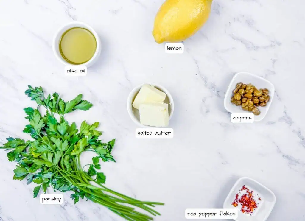 Overhead shot of labeled ingredients for lemon butter sauce: lemon, olive oil, salted butter, capers, red pepper flakes, and flat-leaf parsley.