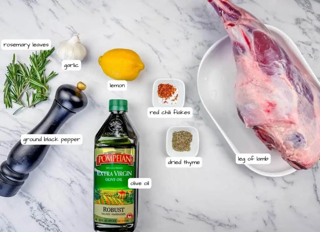 Overhead shot of labeled ingredients for roast leg of lamb, including a leg of lamb, Pompeian olive oil, lemon, garlic, rosemary, thyme, and red chili flakes.