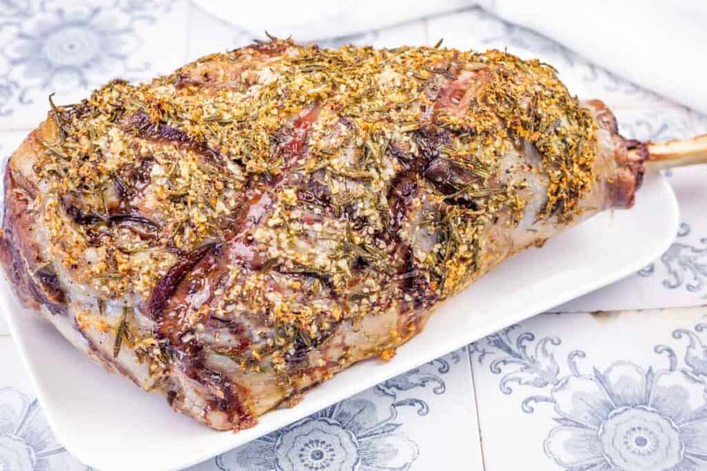 A whole roasted leg of lamb seasoned with herbs on a white serving platter.