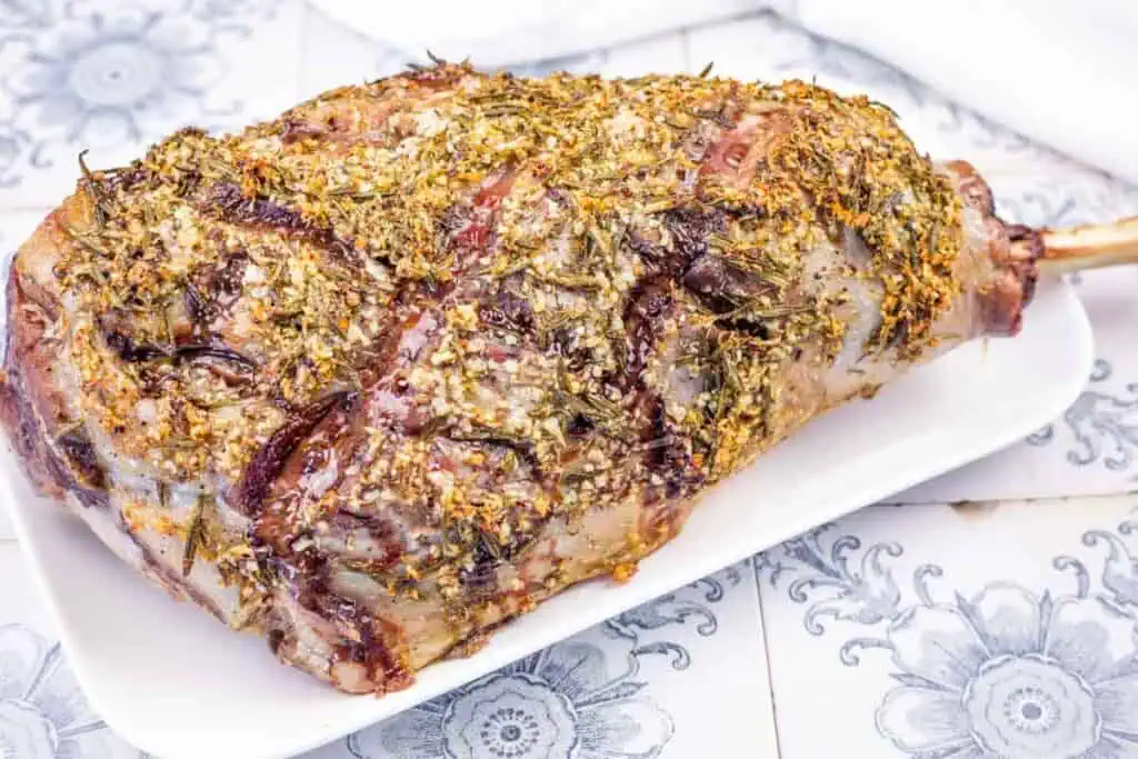 A whole roasted leg of lamb seasoned with herbs on a white serving platter.