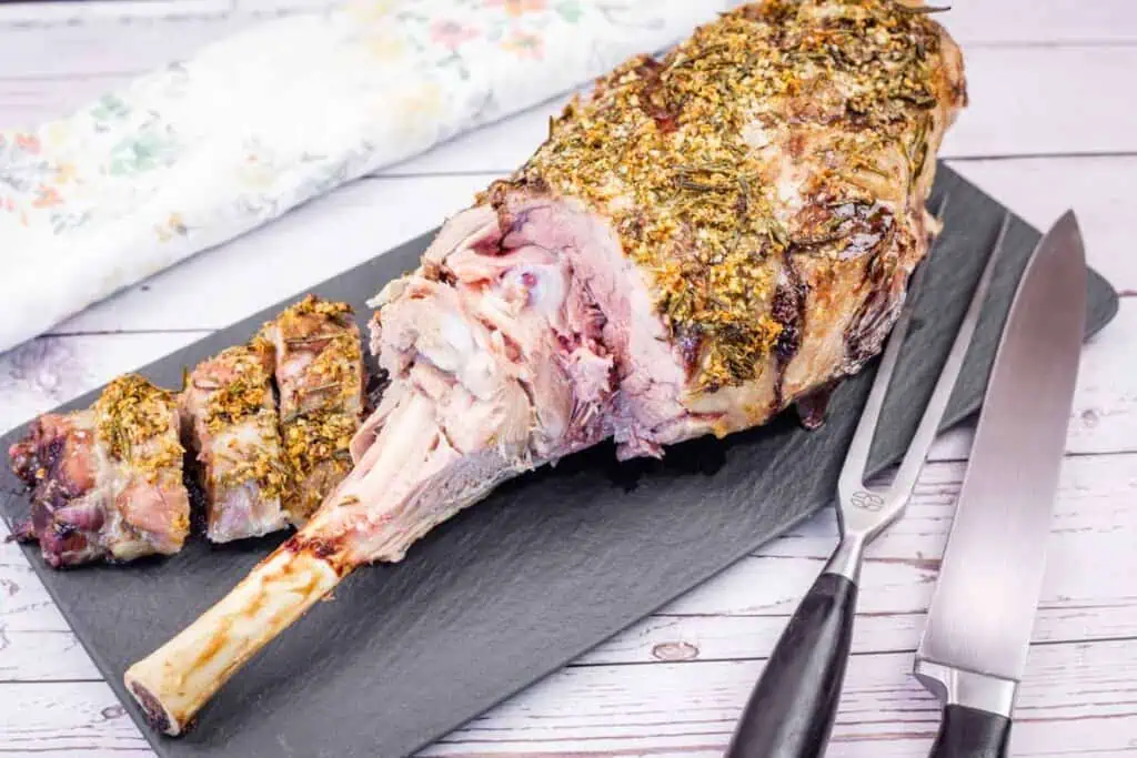 A partially carved roast leg of lamb seasoned with herbs on a black serving platter with carving utensils.