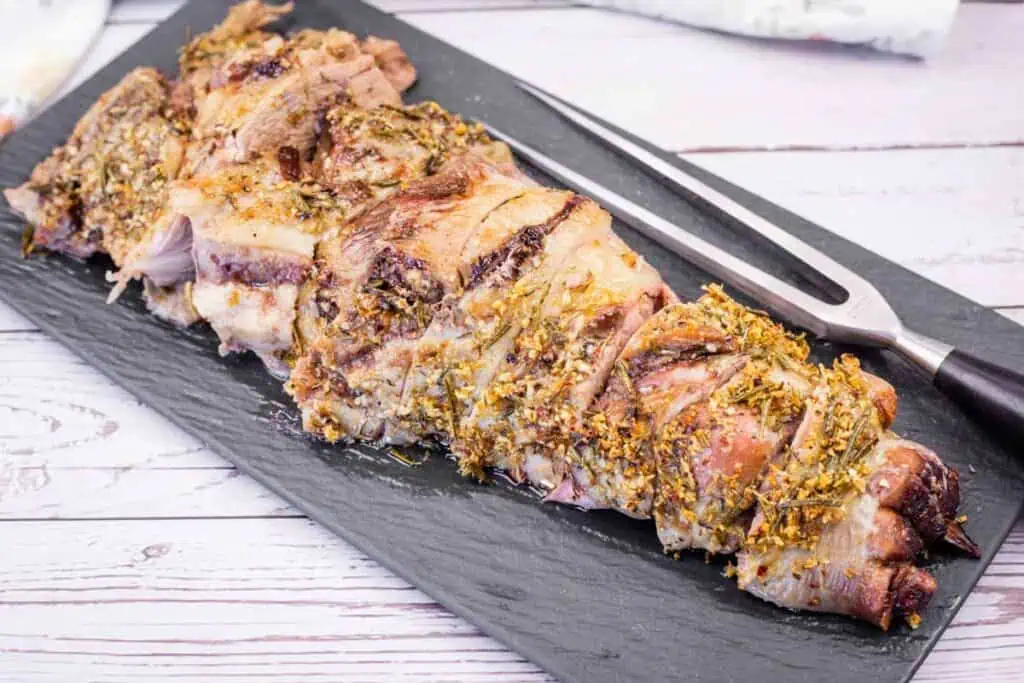 A black platter with sliced, roasted leg of lamb, seasoned with herbs.