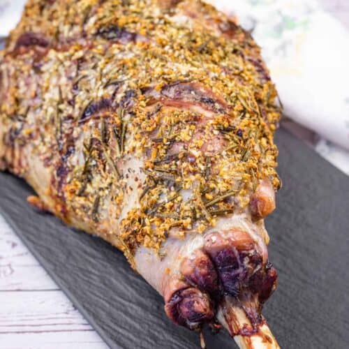 A whole roasted leg of lamb seasoned with herbs on a black serving platter.