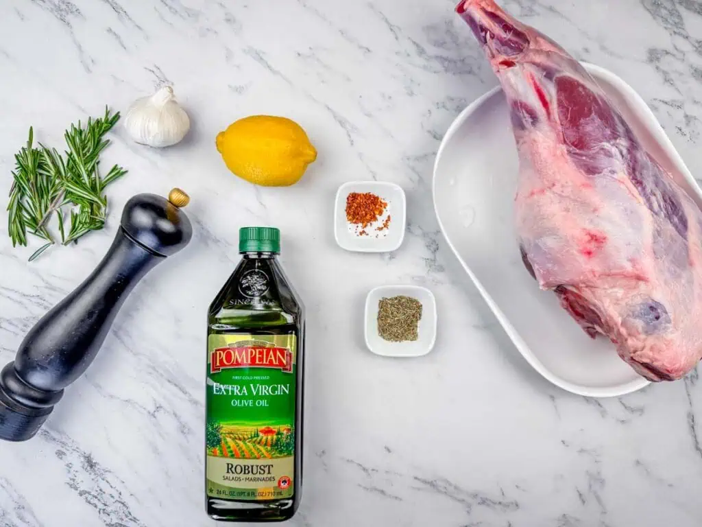 Overhead shot of ingredients for roast leg of lamb, including a leg of lamb, olive oil, lemon, garlic, rosemary, and spices.