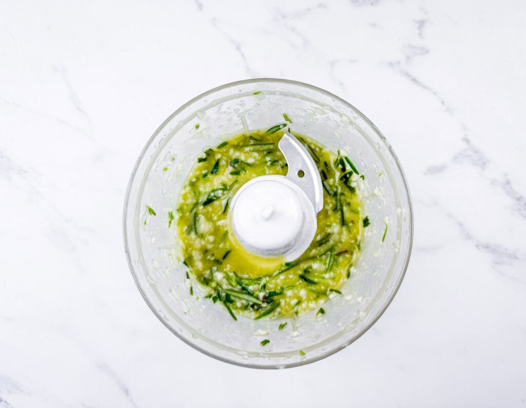 Overhead shot of chopped basil, garlic, and olive oil being processed in a food processor.