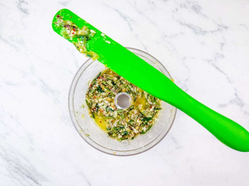 Overhead shot of chopped herbs and spices for chimichurri in a food processor, with a green spatula.