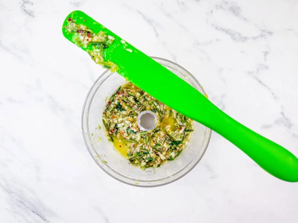 Overhead shot of chopped herbs and spices for chimichurri in a food processor, with a green spatula.