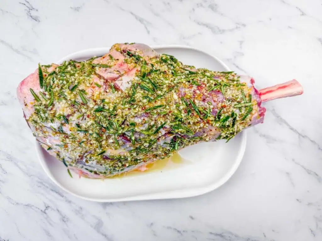 Overhead shot of a raw leg of lamb on a white platter, coated in a herb and garlic marinade.