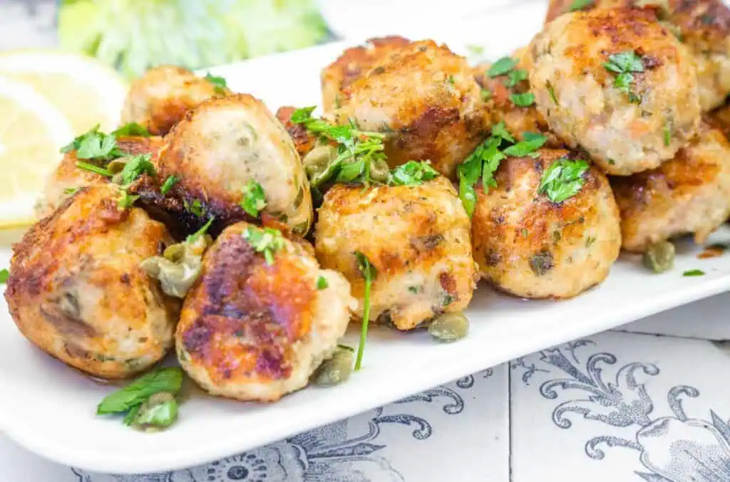 A white platter filled with golden-brown turkey meatballs garnished with capers and fresh parsley, with lemon in the background.