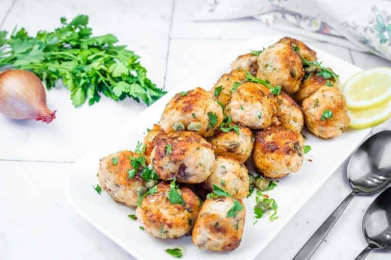 A white plate piled with golden-brown salmon balls garnished with fresh parsley and capers, served with lemon slices.