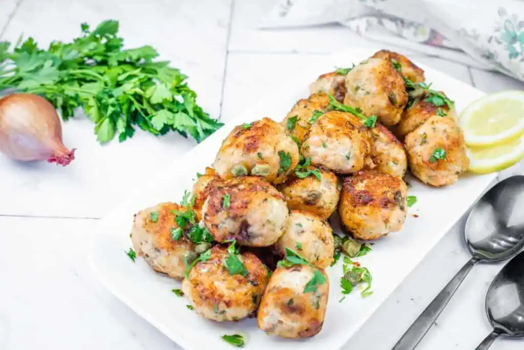 A white platter filled with golden-brown turkey meatballs, garnished with parsley and capers, served with lemon slices and spoons.