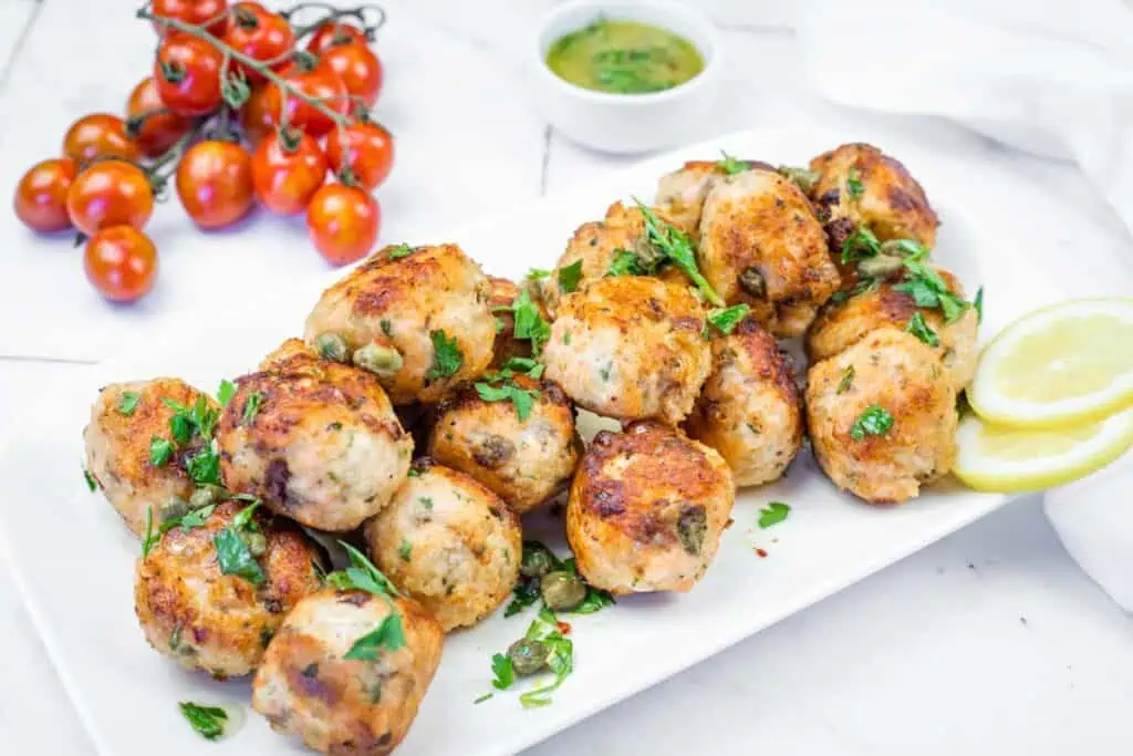 A white platter filled with golden-brown chicken meatballs, garnished with parsley and capers, served with lemon slices and a green sauce.