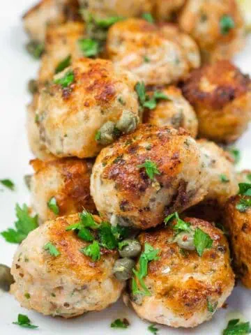 A close-up of a stack of golden-brown meatballs garnished with capers and fresh parsley.