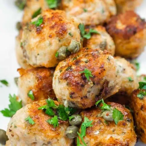 A close-up of a stack of golden-brown meatballs garnished with capers and fresh parsley.