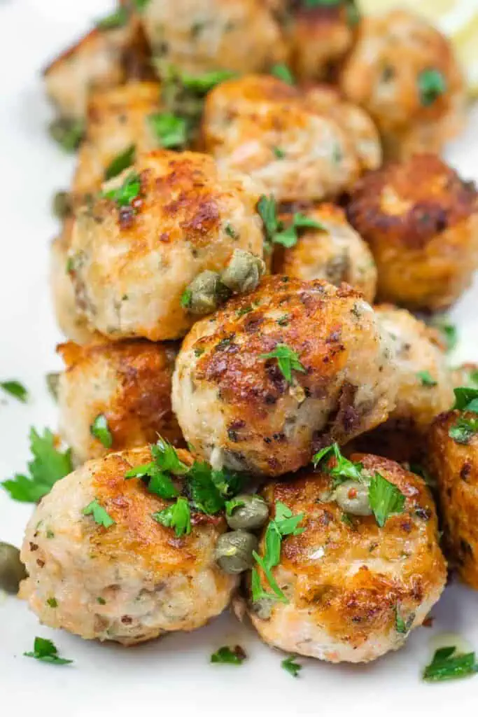 A close-up of a stack of golden-brown meatballs garnished with capers and fresh parsley.