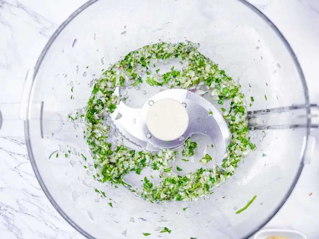 Overhead shot of chopped fresh herbs and shallots being processed in a food processor.