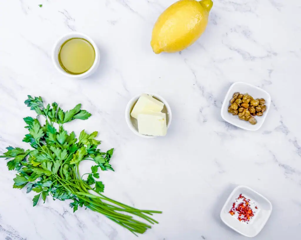 Overhead shot of ingredients for a lemon-caper sauce: lemon, olive oil, butter, capers, red pepper flakes, and parsley.