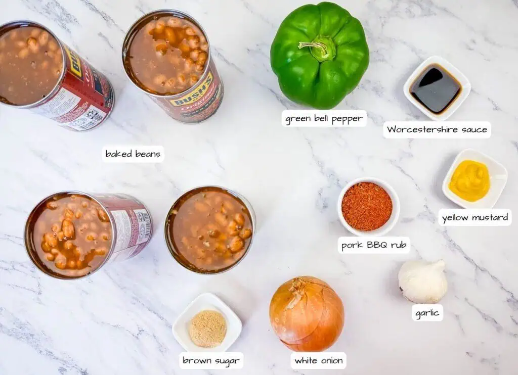 Cans of baked beans, green bell pepper, Worcestershire sauce, yellow mustard, pork BBQ rub, garlic, white onion, and brown sugar arranged on a marble surface, each labeled.