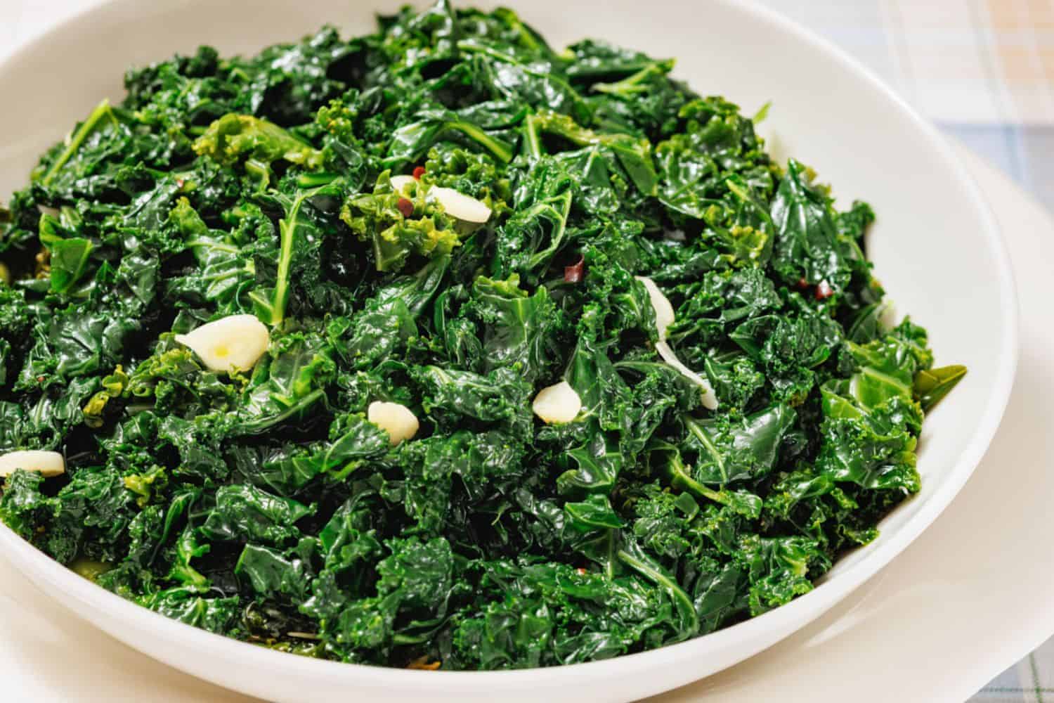 A white bowl filled with sautéed kale and sliced garlic on a light-colored surface.