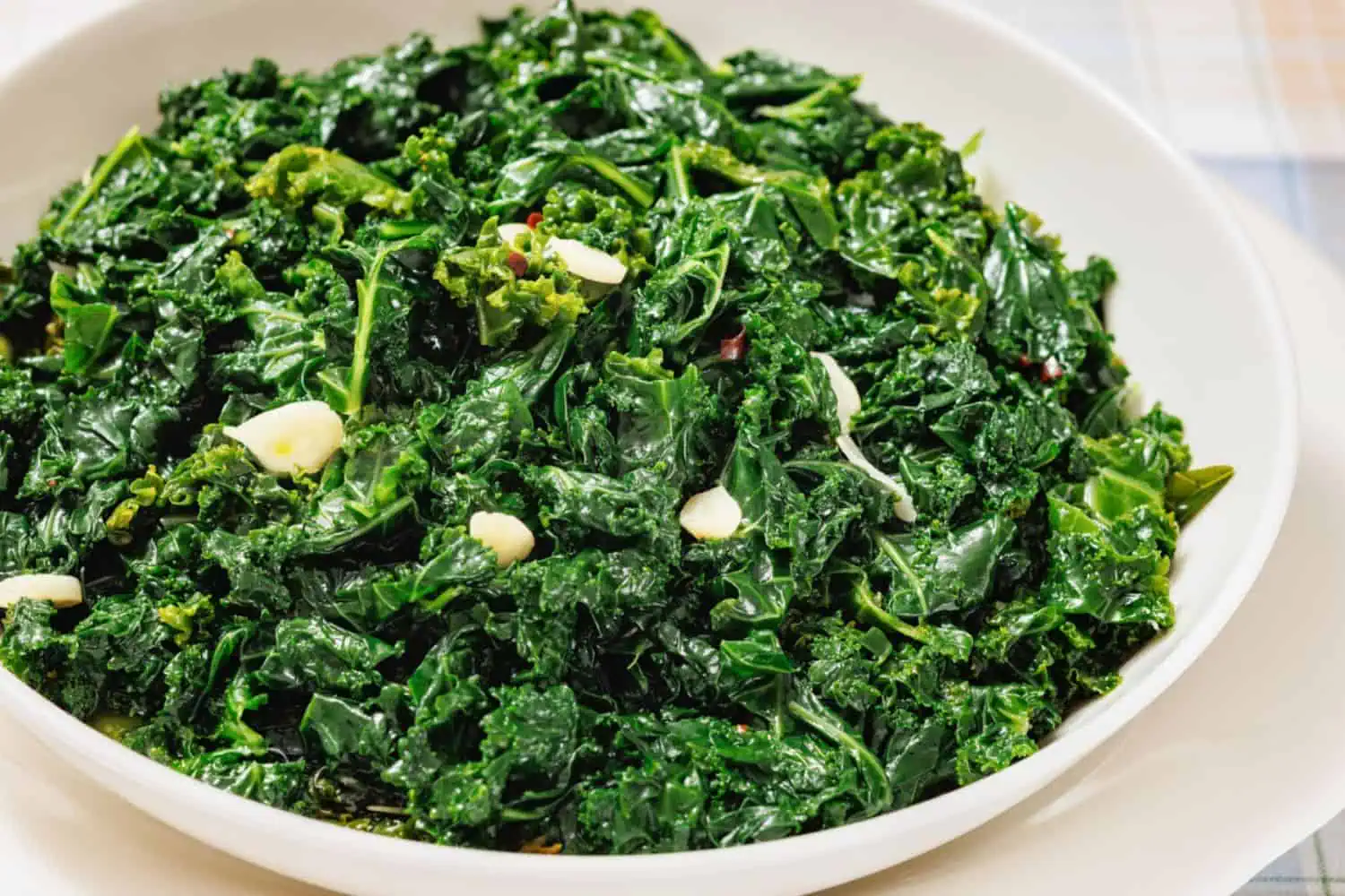 A white bowl filled with sautéed kale and sliced garlic on a light-colored surface.