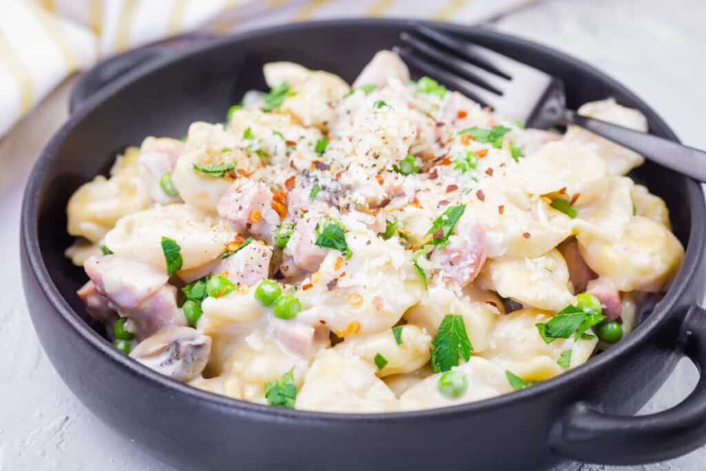 A black bowl filled with Skillet Cheese Tortellini with Ham & Peas, topped with grated cheese and red pepper flakes, with a fork resting on the side.