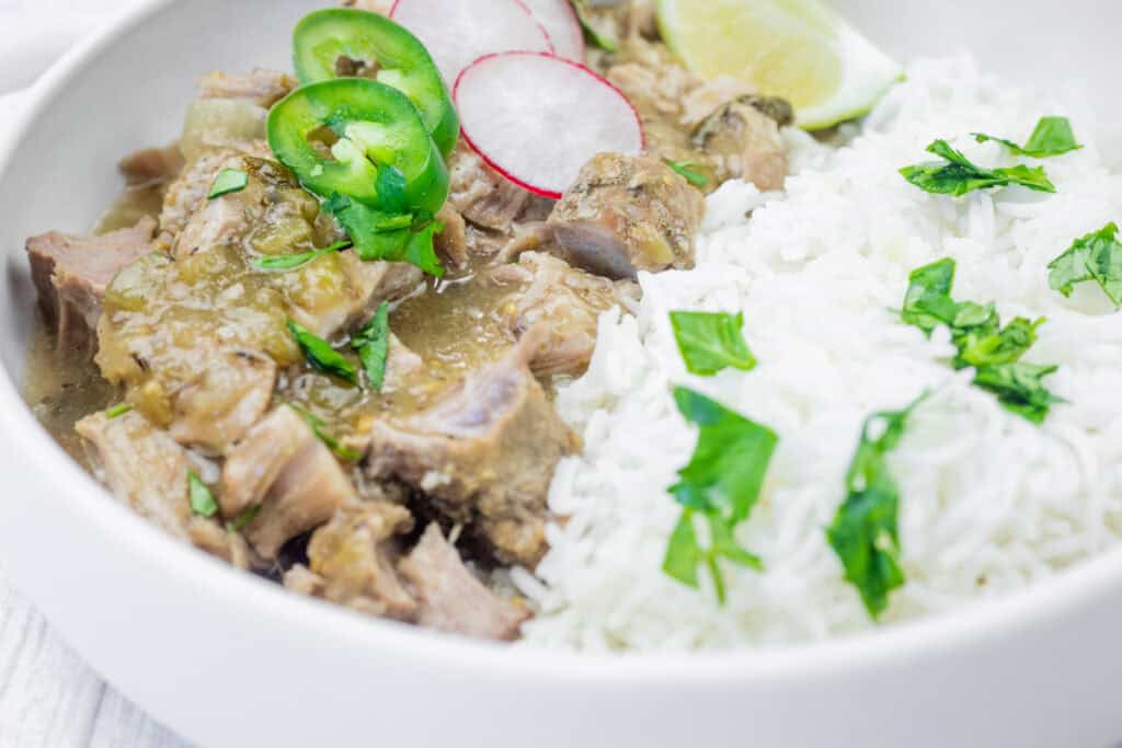 Close-up of a bowl of slow cooker pork chile verde served with white rice, garnished with fresh cilantro, sliced jalape&ntilde;os, radish slices, and a wedge of lime.