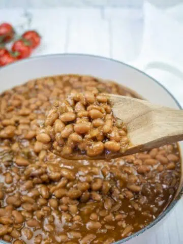 A wooden spoon holding baked beans above a pot filled with baked beans in sauce; tomatoes are visible in the background.