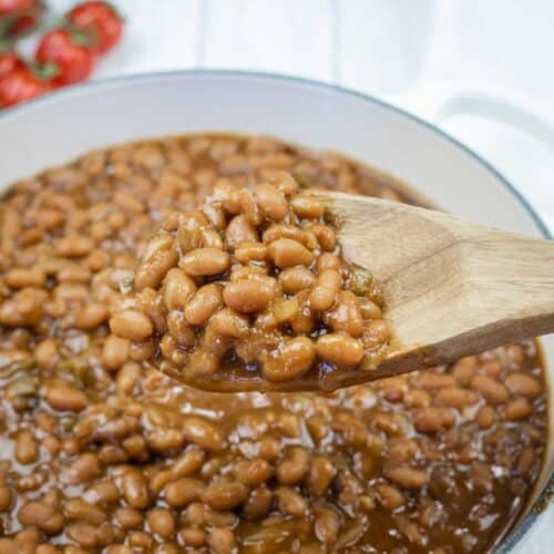 A wooden spoon holding baked beans above a pot filled with baked beans in sauce; tomatoes are visible in the background.