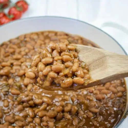 A wooden spoon holding baked beans above a pot filled with baked beans in sauce; tomatoes are visible in the background.