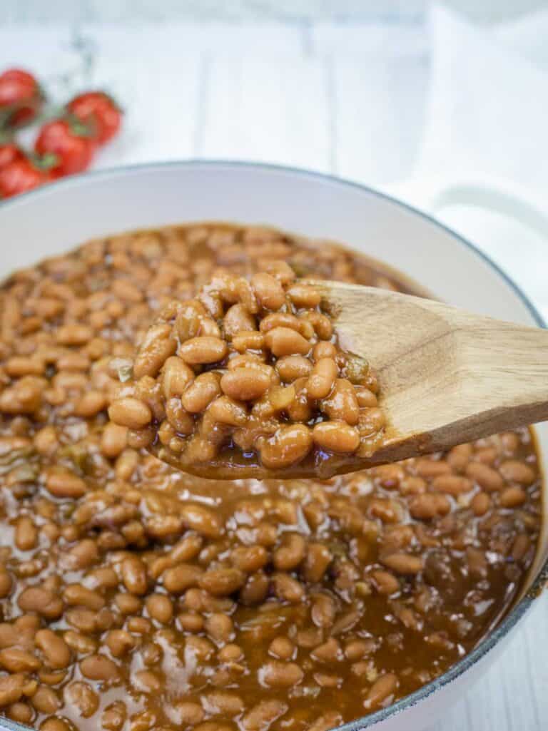 A wooden spoon holding baked beans above a pot filled with baked beans in sauce; tomatoes are visible in the background.