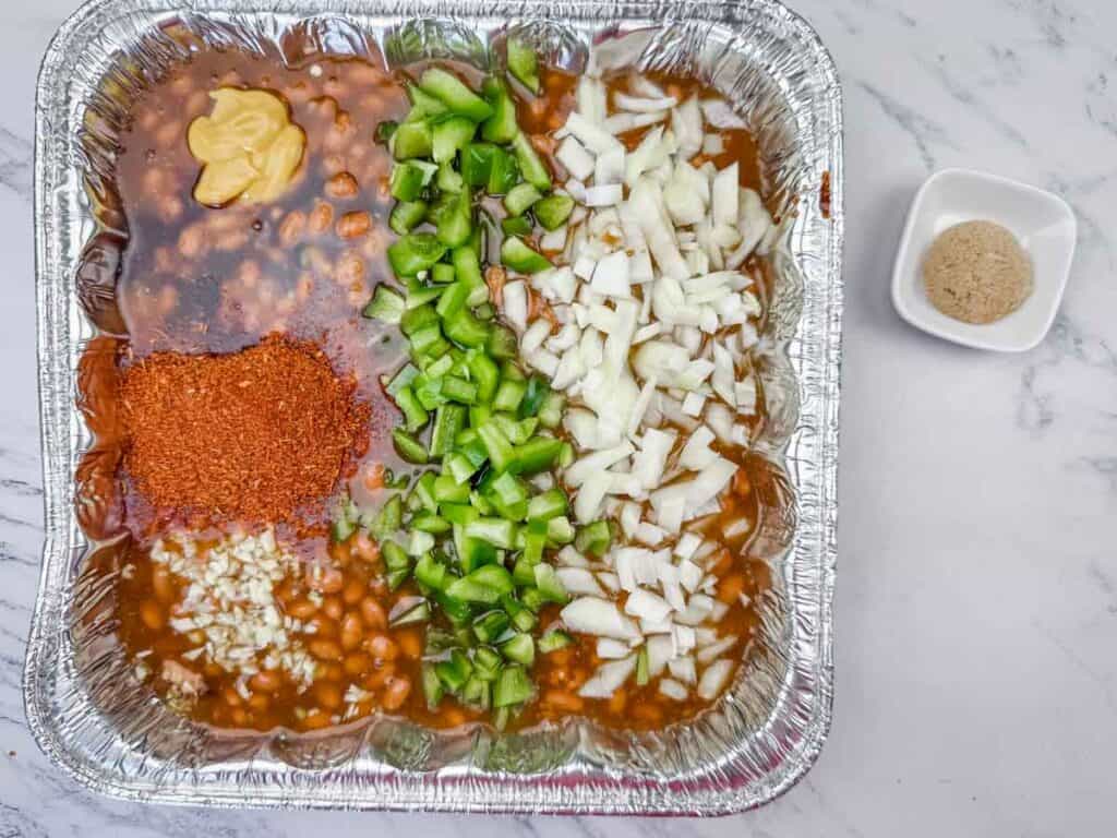 A foil tray with baked beans, chopped green bell peppers, chopped onions, minced garlic, yellow mustard, and chili powder, with a small bowl of brown sugar on the side.