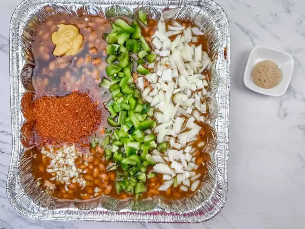 A foil tray with baked beans, chopped green bell peppers, chopped onions, minced garlic, yellow mustard, and chili powder, with a small bowl of brown sugar on the side.