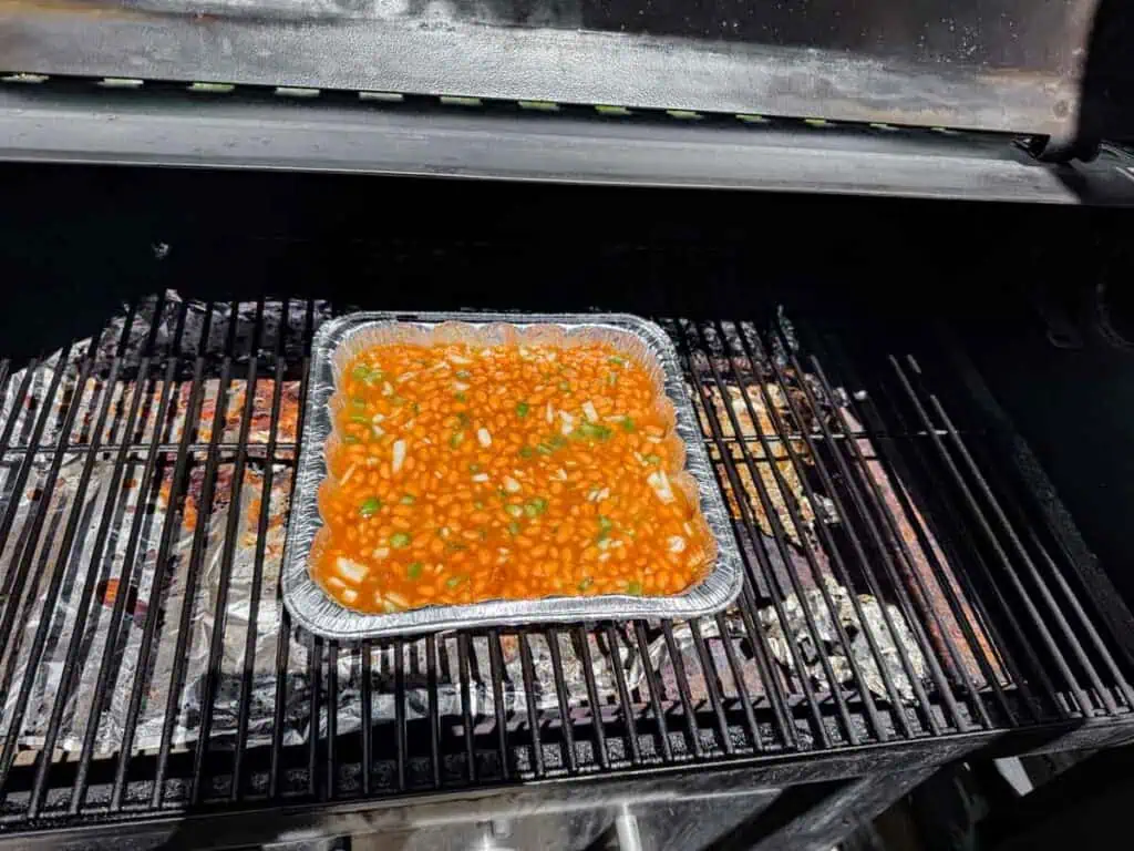 A foil tray filled with baked beans and chopped vegetables sits on a grill inside a barbecue smoker.