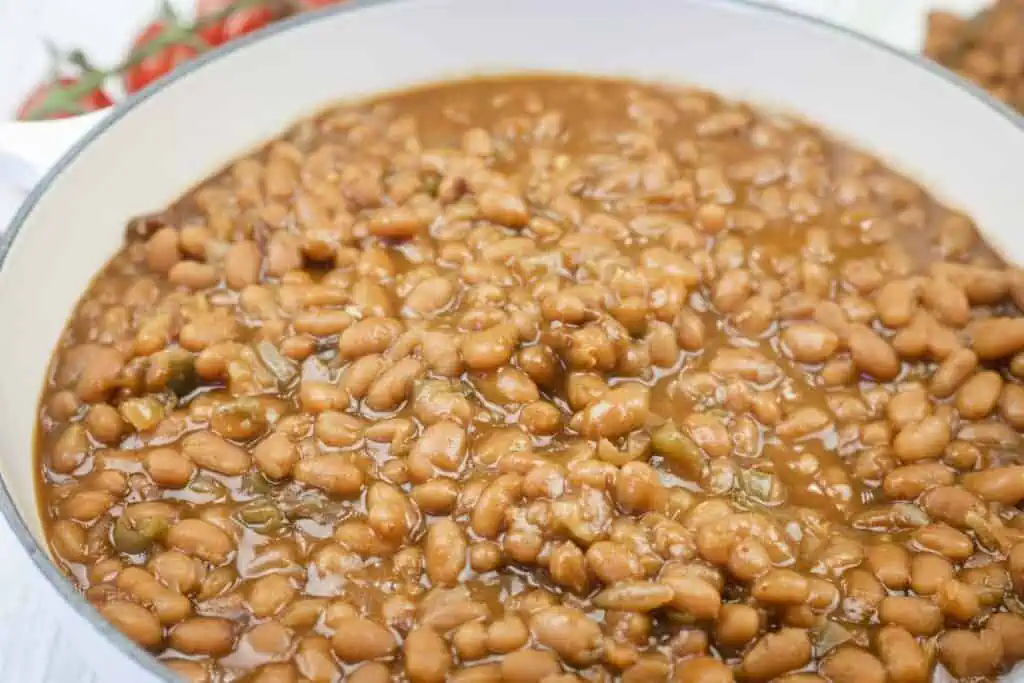 A close-up view of baked beans in a white pot, showing the beans in a thick, brown sauce with visible small pieces of vegetables.
