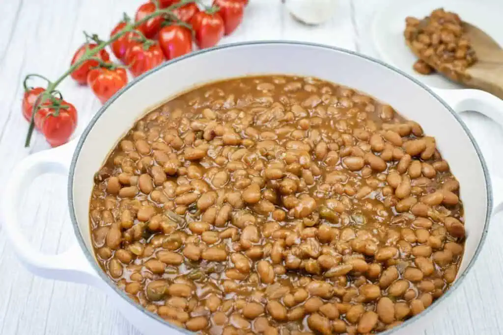 A white pot filled with baked beans in sauce sits on a white surface, with fresh tomatoes and a spoon with beans in the background.