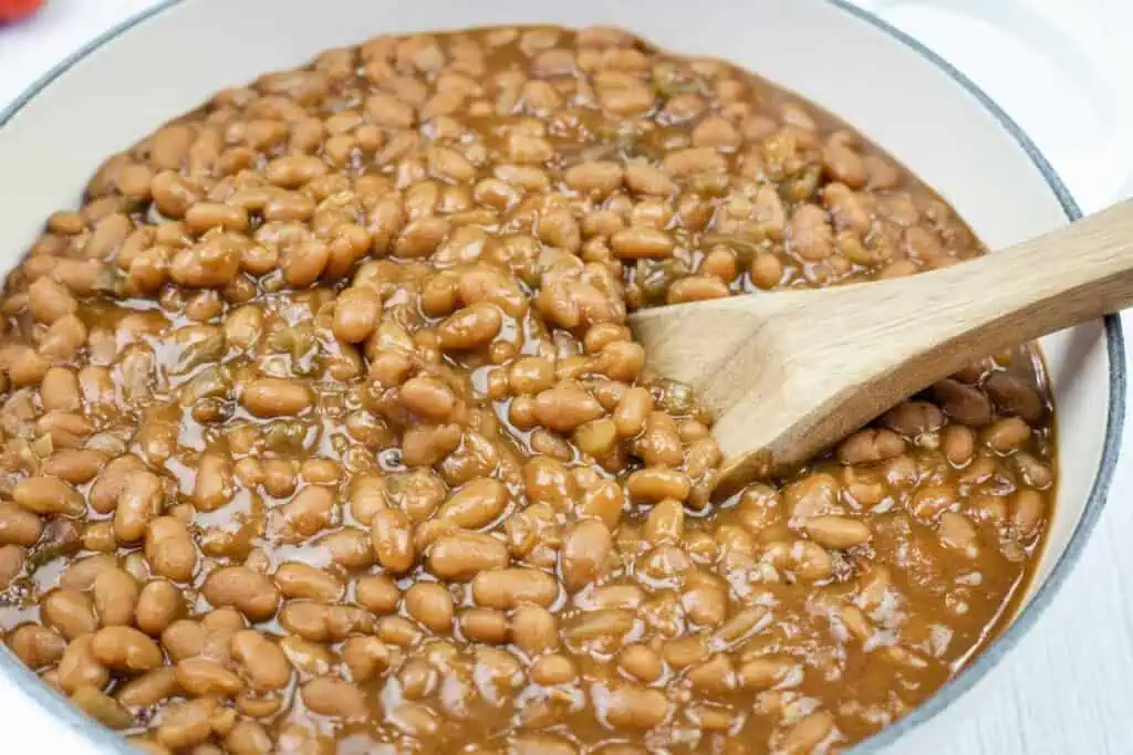A pot of baked beans in sauce with a wooden spoon stirring them.