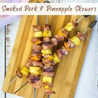 Three metal skewers with pieces of smoked pork, pineapple, red onion, and yellow bell pepper on a wooden cutting board. Text reads "Smoked Pork & Pineapple Skewers.