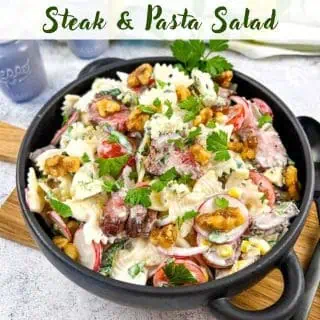 A black bowl filled with a steak & pasta salad containing farfalle, steak slices, radishes, cherry tomatoes, lettuce, nuts, and crumbled blue cheese, garnished with parsley.