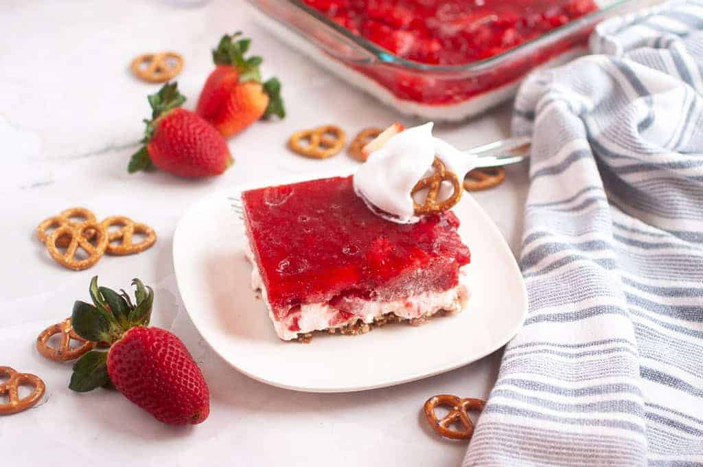 A slice of Strawberry Pretzel Salad topped with whipped cream and a pretzel sits on a white plate.