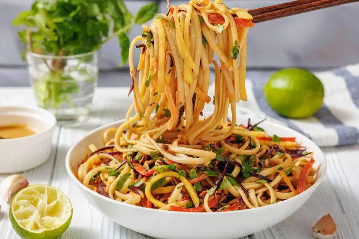 A bowl of Thai Noodle Salad with Mango and Spicy Peanut Dressing is lifted by chopsticks.
