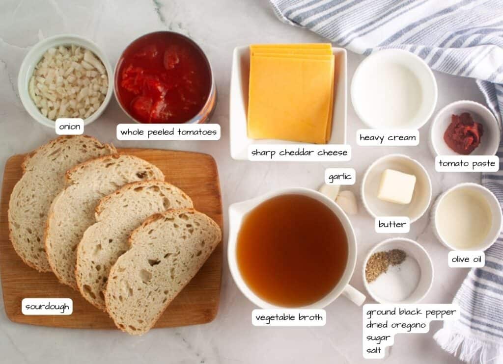 Ingredients for a recipe laid out on a table, including sliced sourdough bread, onion, whole peeled tomatoes, cheddar cheese, heavy cream, tomato paste, garlic, broth, butter, olive oil, and seasonings.
