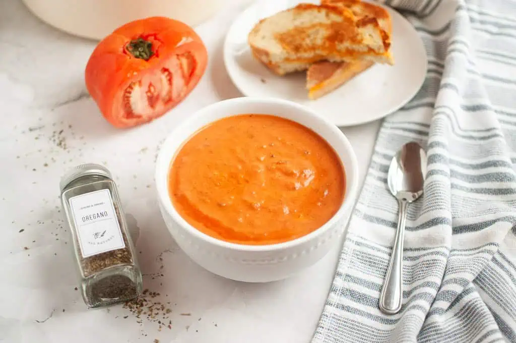 A bowl of creamy tomato soup with a spoon, served with a grilled cheese sandwich and a fresh tomato.