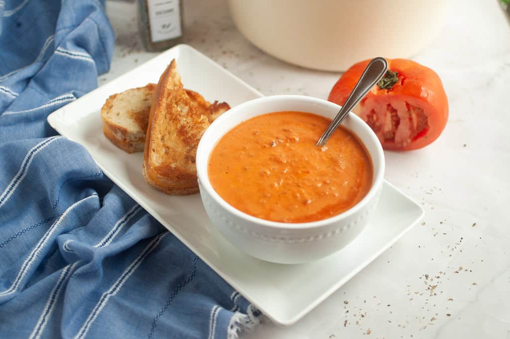 A bowl of creamy tomato soup with a spoon, served with a grilled cheese sandwich and a fresh tomato.