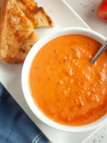 A white bowl of creamy orange-red tomato soup with a spoon, served with a grilled cheese sandwich.