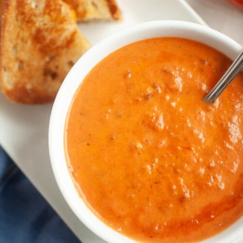 A white bowl of creamy orange-red tomato soup with a spoon, served with a grilled cheese sandwich.