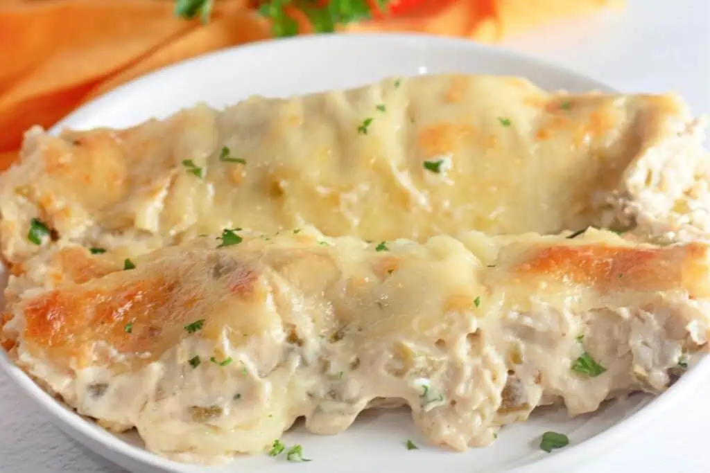 Two creamy, cheese-topped chicken enchiladas garnished with small pieces of parsley on a white plate.