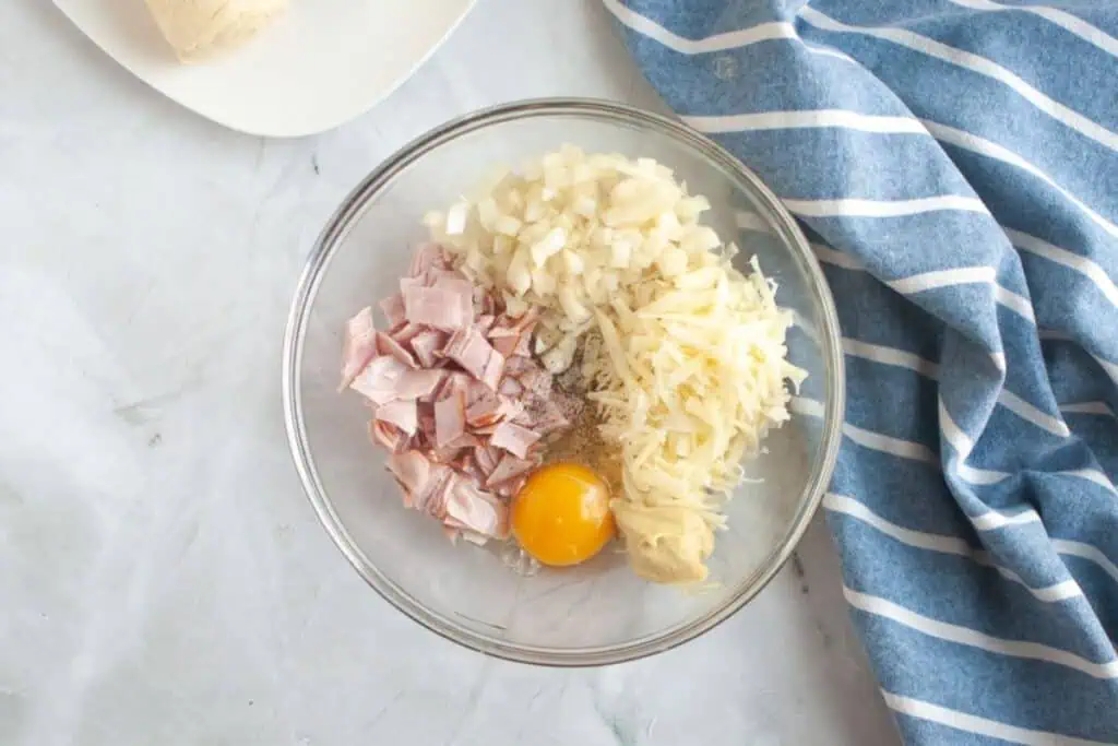 Overhead shot of a glass bowl containing diced ham, shredded cheese, diced onion, an egg, mustard, and pepper.