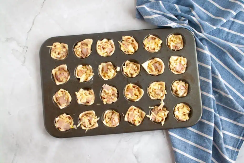 Overhead shot of a muffin tin filled with small pastry cups containing a mixture of ham and cheese.